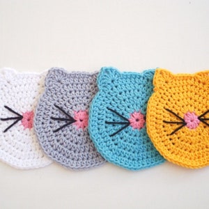 May include: Four crocheted cat-shaped coasters in white, gray, blue, and yellow. Each coaster has a pink nose and black whiskers.