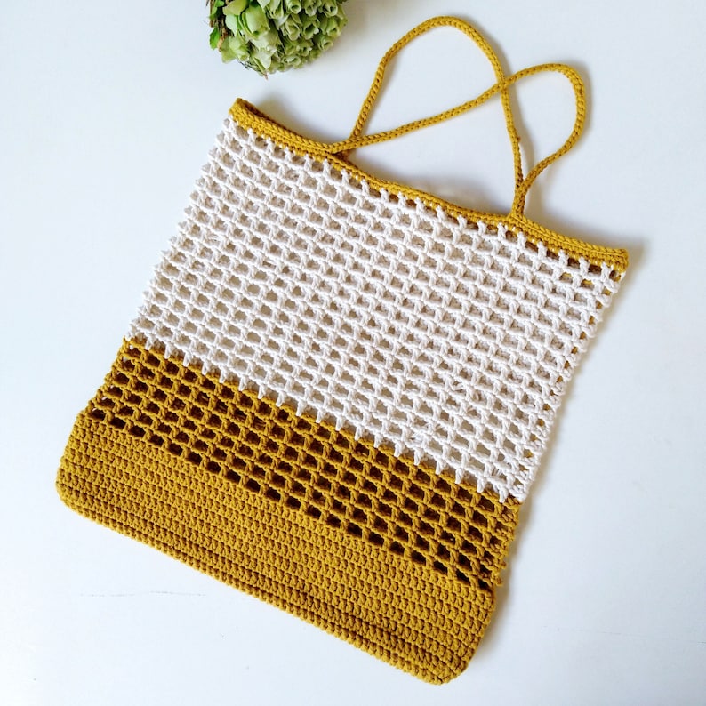 May include: A crocheted tote bag with a white and mustard yellow color scheme. The bag has a wide, flat base and a single handle.