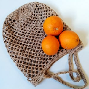 May include: A crocheted beige shopping bag with three bright orange oranges inside. The bag has a net-like design and a handle. The oranges are round and have a smooth texture. The bag is made of a soft, natural fiber.