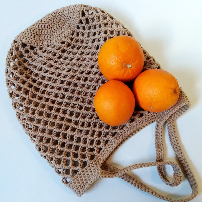 May include: A crocheted beige shopping bag with three bright orange oranges inside. The bag has a net-like design and a handle. The oranges are round and have a smooth texture. The bag is made of a soft, natural fiber.