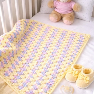 Puede incluir: Una manta de bebé hecha a mano con un patrón de cuadrados de la abuela, con colores amarillo, lavanda y azul claro. La manta se combina con botines de bebé amarillos y un chupete. Un oso de peluche está en el fondo.