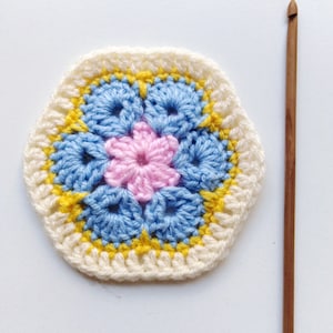 May include: A crocheted hexagon with a pink flower in the center, surrounded by blue and yellow flowers. The hexagon is made with white yarn and has a yellow border.