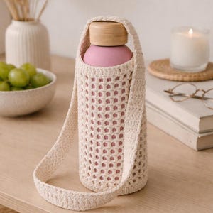 May include: A pink water bottle with a wooden cap is held in a cream-coloured, open-weave crochet carrier. The carrier has a long strap for easy carrying. The bottle sits on a light-coloured wooden surface.