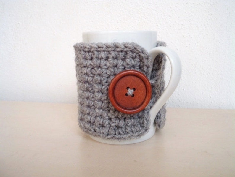 May include: A gray crocheted mug cozy with a wooden button. The cozy is wrapped around a white ceramic mug.