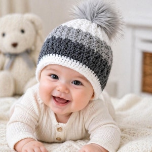 May include: A baby wearing a handmade crochet hat with gray, white, and dark gray stripes and a gray faux fur pom-pom. The baby is smiling and wearing a cream-colored onesie. A stuffed teddy bear is in the background.