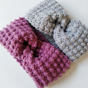 May include: Two crocheted headbands, one in a light gray and one in a deep purple, are tied in a knot. The headbands are made with a textured stitch that gives them a bumpy appearance.