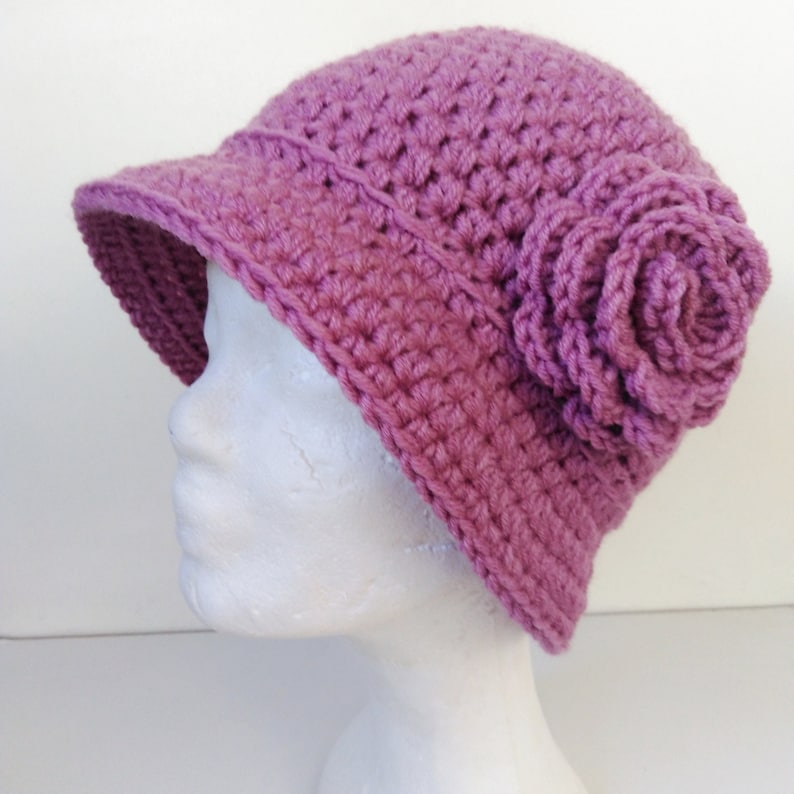 May include: A crocheted cloche hat in a dusty rose color. The hat features a small brim and a large, decorative rose on the side. The hat is made of a textured yarn, and is displayed on a white mannequin head.