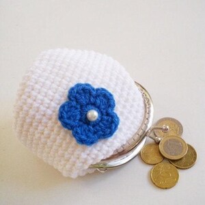 May include: A white crocheted coin purse with a silver metal frame and a blue crocheted flower embellishment featuring a central white pearl bead. Several gold-colored coins are positioned next to the open purse on a plain white surface.