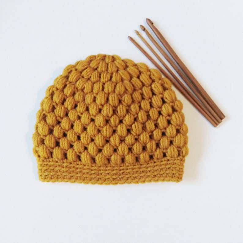 May include: A mustard yellow crocheted beanie hat with a textured bobble stitch pattern. The hat is lying on a white surface with four wooden crochet hooks.