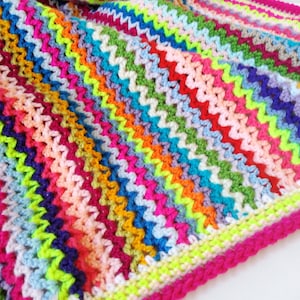 Pode incluir: Uma manta de crochê colorida com um padrão em ziguezague em tons de rosa, azul, verde, amarelo, laranja e roxo. A manta tem uma borda rosa.