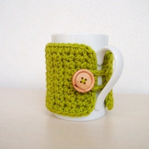 May include: A white ceramic mug with a green crocheted cosy. The cosy has a wooden button with a four-point star design.