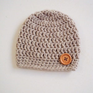 May include: A light beige crochet beanie hat made from thick yarn with a rounded crown and a straight bottom edge. A single, round, light brown wooden button with four holes is attached to the lower right side of the textured knit fabric.