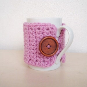 May include: A white ceramic mug with a pink crocheted cozy and a brown wooden button. The cozy is crocheted in a simple stitch pattern.