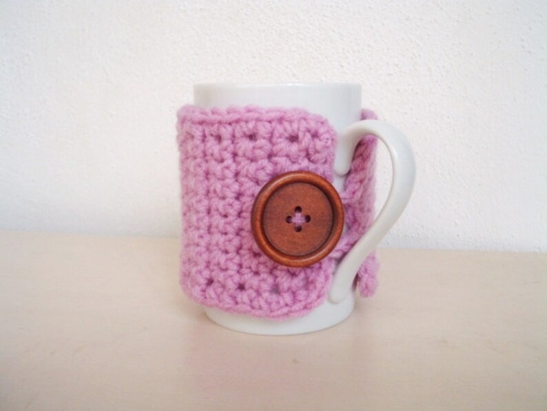 May include: A white ceramic mug with a pink crocheted cozy and a brown wooden button. The cozy is crocheted in a simple stitch pattern.