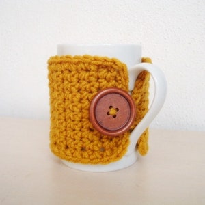 May include: A white ceramic mug with a crocheted mustard yellow cozy. The cozy has a brown wooden button with a flower design.