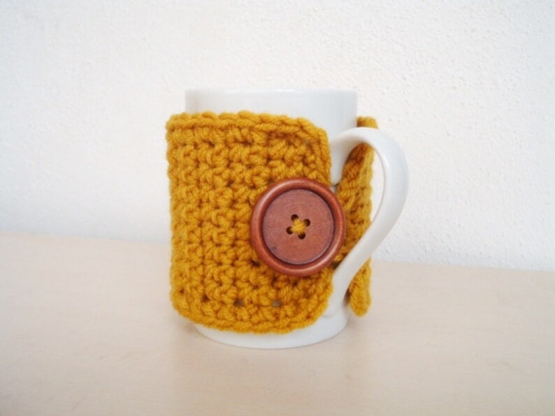 May include: A white ceramic mug with a crocheted mustard yellow cozy. The cozy has a brown wooden button with a flower design.