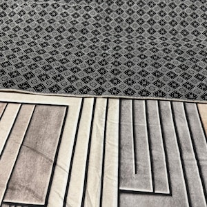 Op de afbeelding: Een zwart-wit gestreept tapijt met een geometrisch ontwerp. Het tapijt heeft een zwarte rand en een beige en grijs geometrisch patroon in het midden.