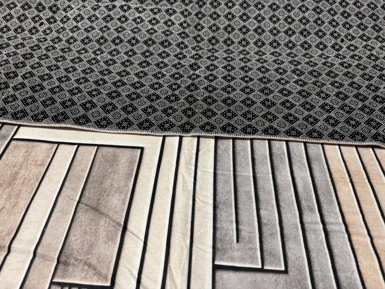 Op de afbeelding: Een zwart-wit gestreept tapijt met een geometrisch ontwerp. Het tapijt heeft een zwarte rand en een beige en grijs geometrisch patroon in het midden.