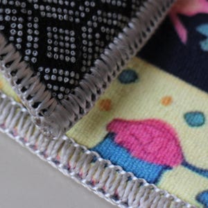 Op de afbeelding: Close-up van de rand van twee stoffen vloerkleden. Het bovenste kleed is zwart met een wit stippenpatroon en een zilvergrijze rand. Het onderste kleed is geel met een roze en blauwe cupcake-ontwerp en een zilvergrijze rand.