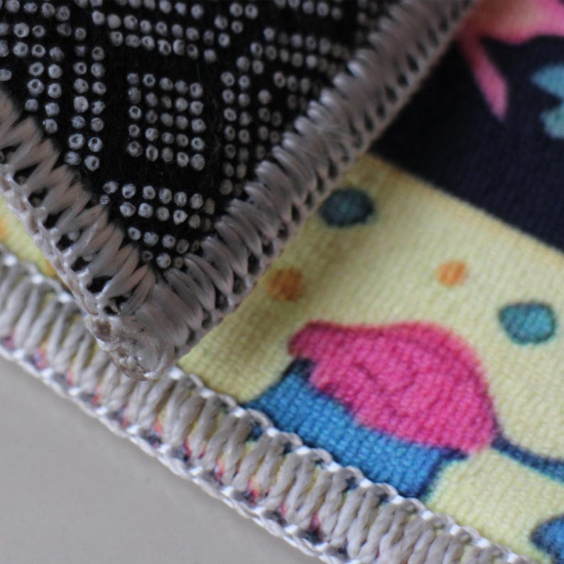 Op de afbeelding: Close-up van de rand van twee stoffen vloerkleden. Het bovenste kleed is zwart met een wit stippenpatroon en een zilvergrijze rand. Het onderste kleed is geel met een roze en blauwe cupcake-ontwerp en een zilvergrijze rand.
