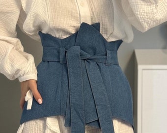 May include: A white long-sleeved blouse with a textured fabric, button-down front, and ruffled shoulder details, paired with a wide blue denim corset-style belt that features a front tie closure with long fabric ends hanging down the front.