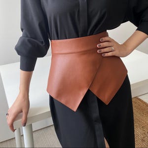 May include: A brown leather wrap belt with an asymmetrical design. The belt is worn over a black dress, creating a layered look. The belt's color is a warm, medium brown, and it appears to be made of smooth leather.