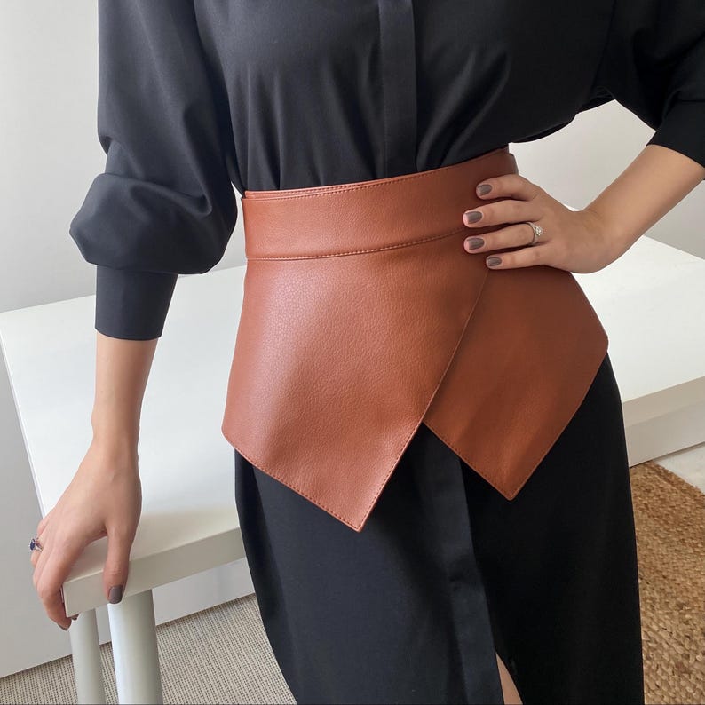 May include: A brown leather wrap belt with an asymmetrical design. The belt is worn over a black dress, creating a layered look. The belt's color is a warm, medium brown, and it appears to be made of smooth leather.