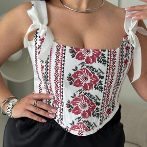 May include: A white corset top featuring a red and black cross-stitch floral pattern and vertical geometric borders. The garment has a square neckline and shoulder straps that tie into bows at the top, with a structured bodice design.