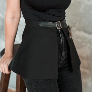 May include: Black waist belt with a flared skirt design. The belt features a rectangular buckle and a leather strap. A small decorative element is attached to the side. The belt is worn over black jeans.
