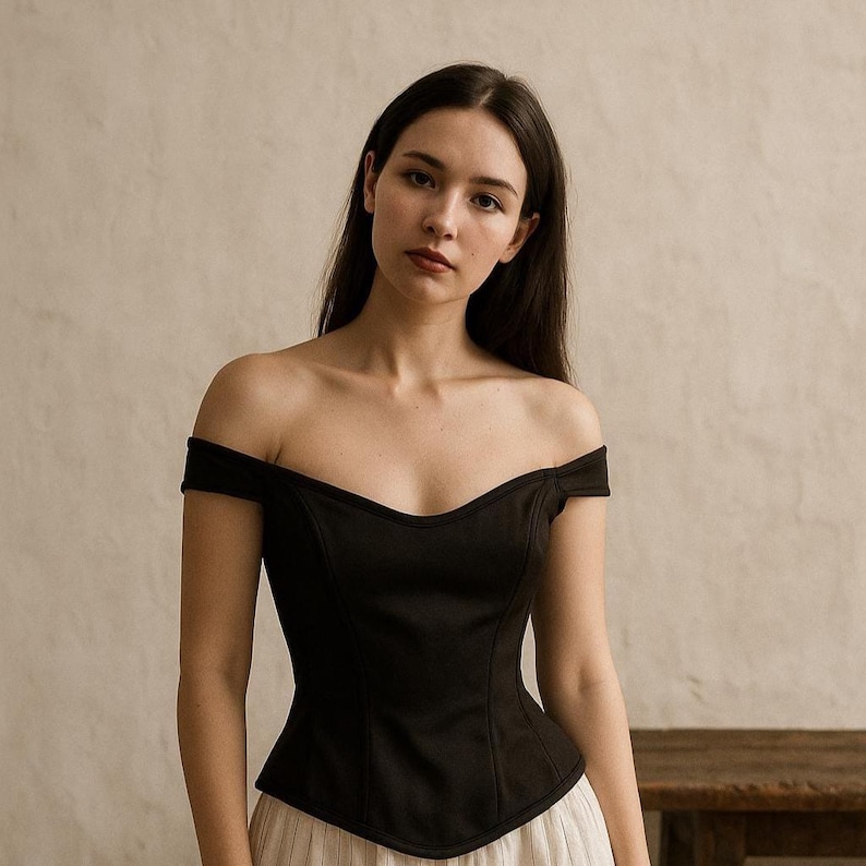 May include: A black off-the-shoulder top with a sweetheart neckline. The top has short sleeves and a fitted bodice. The model is wearing a cream-colored skirt. The background is a neutral color.