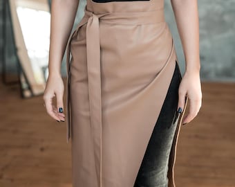 Puede incluir: Una falda cruzada de piel sintética en color beige con diseño de cintura alta y un cinturón de lazo a juego. La falda tiene un acabado liso y mate con una superposición frontal diagonal que crea una abertura lateral, sobre pantalones grises.