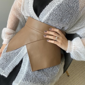 May include: A tan-colored leather belt with an asymmetrical design. The belt wraps around the waist and features a wide, overlapping front panel. The belt is a neutral color and appears to be made of smooth leather. The belt is a fashion accessory.