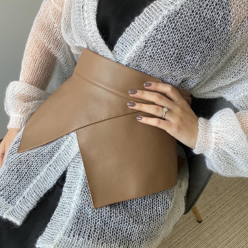 May include: A tan-colored leather belt with an asymmetrical design. The belt wraps around the waist and features a wide, overlapping front panel. The belt is a neutral color and appears to be made of smooth leather. The belt is a fashion accessory.