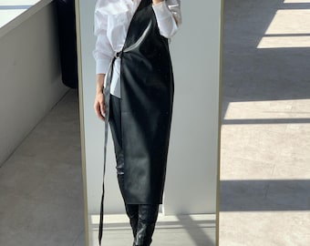 Puede incluir: Una camisa blanca de manga larga con botones debajo de un vestido tipo delantal cruzado de piel sintética negra. El vestido cuenta con un cierre de hebilla lateral con una tira larga colgante, combinado con pantalones negros y botas negras.