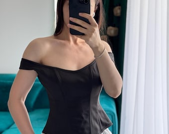 May include: Black off-the-shoulder corset top with a sweetheart neckline. The top has a fitted bodice and is paired with white and light-colored striped pants. The top is made of a smooth, solid fabric.