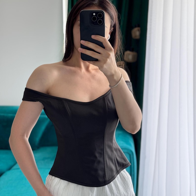 May include: Black off-the-shoulder corset top. The top has a sweetheart neckline and a fitted bodice. The garment is paired with white and light-colored striped pants. The top is made of a smooth, solid fabric.