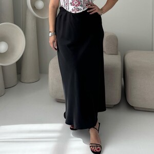 May include: A black maxi skirt with a high waist. The skirt is paired with a white top with a floral pattern. The outfit is completed with black sandals. The skirt is made of a smooth, flowing fabric.