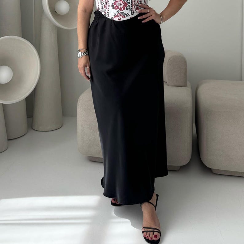 May include: A black maxi skirt with a high waist. The skirt is paired with a white top with a floral pattern. The outfit is completed with black sandals. The skirt is made of a smooth, flowing fabric.