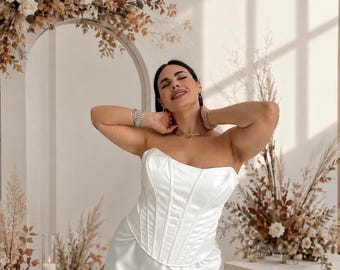 May include: A strapless white corset top featuring vertical boning seams and a smooth satin finish. The bodice has a structured, fitted silhouette with a sweetheart neckline, crafted from a lustrous white fabric that reflects light.