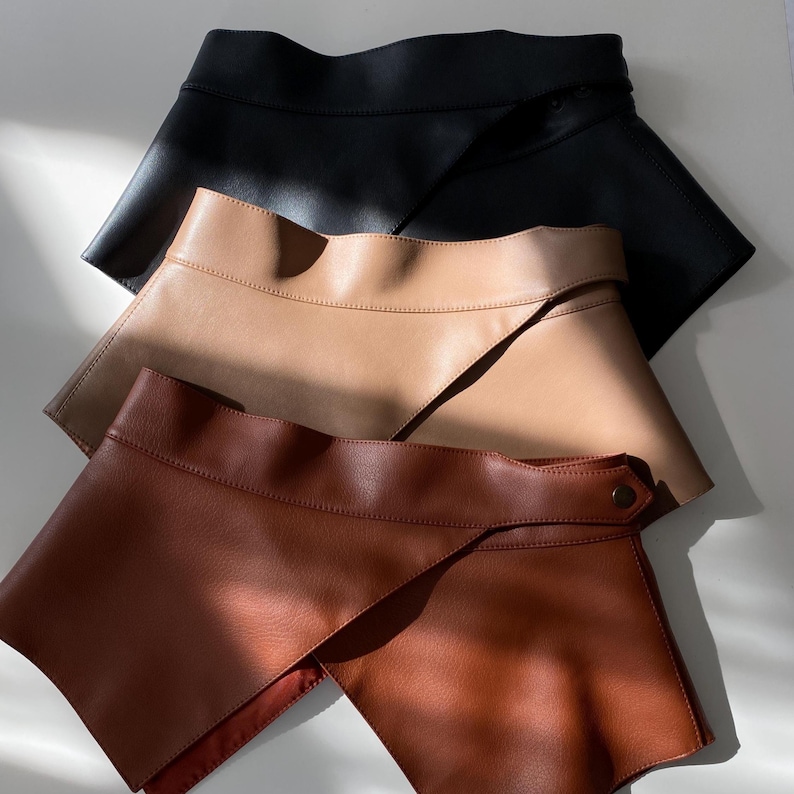 May include: Three leatherette skirts in black, tan, and brown. Each skirt has a unique asymmetrical design with a wrap-around style. The skirts are displayed on a white surface, showcasing their different colors and textures.