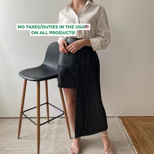 May include: A crisp white long-sleeved button-down shirt paired with a black skirt featuring a smooth leather-like panel on one side and a long, vertically pleated fabric panel on the other. The text NO TAXES/DUTIES IN THE USA ON ALL PRODUCTS! is visible.