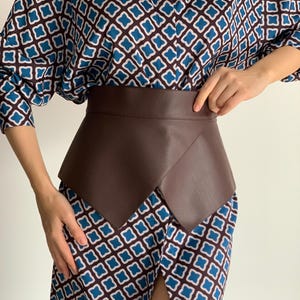 May include: A brown leather belt with a unique, angular design. The belt is a rich, dark brown color and appears to be made of smooth leather. The belt is worn over a patterned dress with blue and white geometric shapes.
