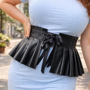Peut inclure: Ceinture noire en similicuir avec un design de jupe plissée. La ceinture présente une ceinture structurée avec une fermeture à nouer en ruban de satin. La partie jupe est plissée, créant une silhouette évasée. La ceinture est portée sur une robe bleu clair.