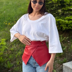 May include: A red leather belt with a unique asymmetrical design. The belt features a button closure and is worn over blue jeans and a white t-shirt. The belt's vibrant color adds a bold accent to the outfit.