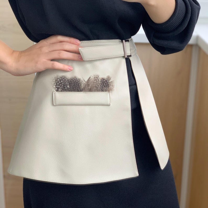 May include: Cream-colored leather belt with a rectangular pocket adorned with feathers. The belt features a silver-tone buckle and a flap closure. The design is a unique fashion accessory.