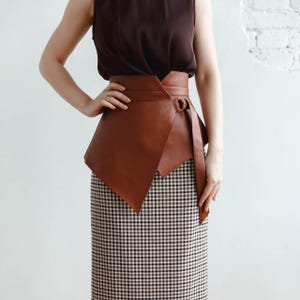 May include: A brown leather wrap belt with a D-ring buckle, worn over a checkered skirt. The belt has an asymmetrical design with a long, flowing strap. The skirt features a black, white, and brown plaid pattern.