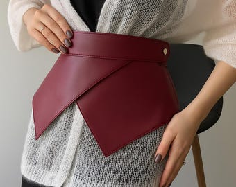 May include: A wide, deep burgundy faux leather corset-style belt featuring an asymmetrical wrap-around design with pointed front panels and a single metallic stud closure on the side, worn over a white sheer knit cardigan and black top.