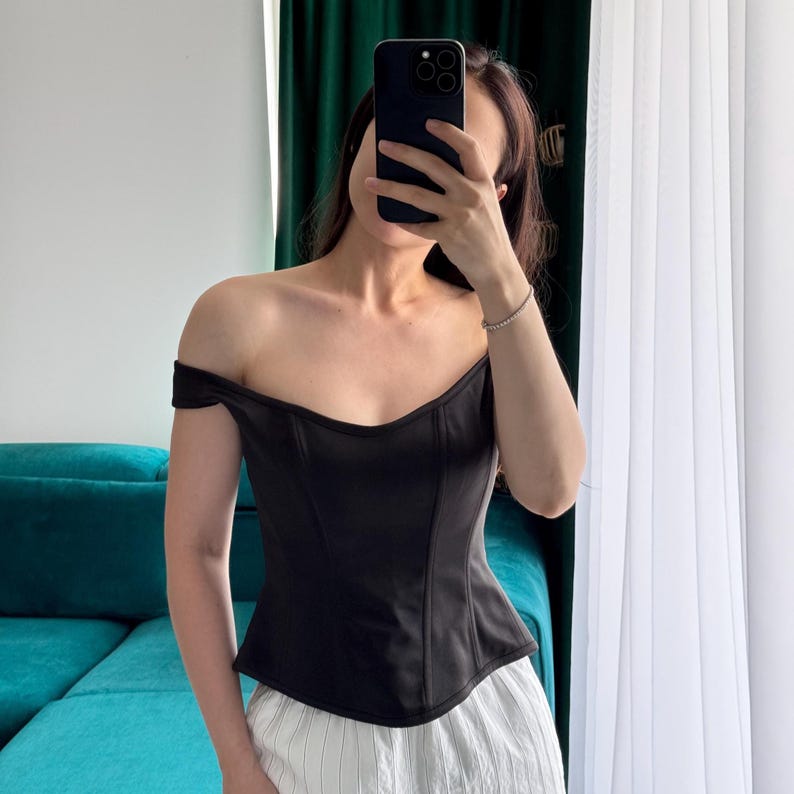 May include: Black off-the-shoulder corset top. The top has a sweetheart neckline and a fitted silhouette. The fabric appears to be a smooth, matte material. The top is paired with white and light-colored striped pants.