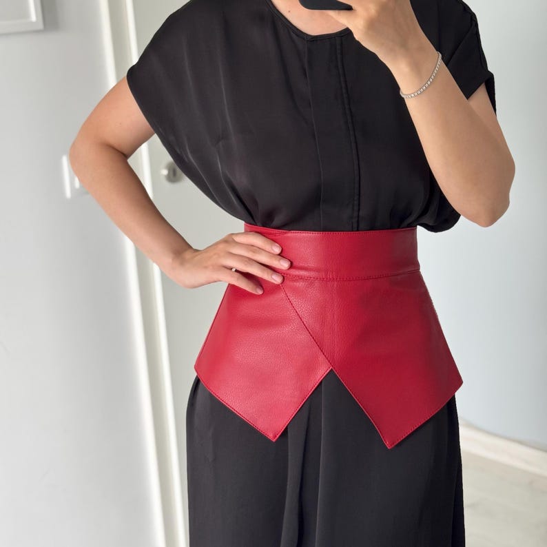 May include: A bright red leather belt with a unique geometric design. The belt is wide and wraps around the waist, creating a bold and stylish look. The belt is worn over a black dress.