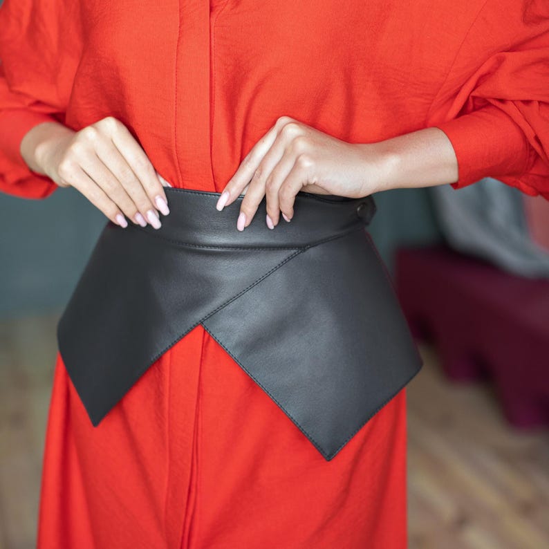 May include: A black leather waist belt with a geometric design. The belt is worn over a bright red dress. The belt has a wide, angular front panel and a narrow back strap. The person is adjusting the belt.